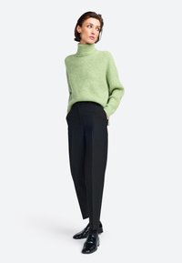 Grüner, strukturierter Rollkragenpullover kombiniert mit schwarzen maßgeschneiderten Hosen und glänzenden schwarzen Slippern. Schlichte, klare Linien dominieren das Outfit.