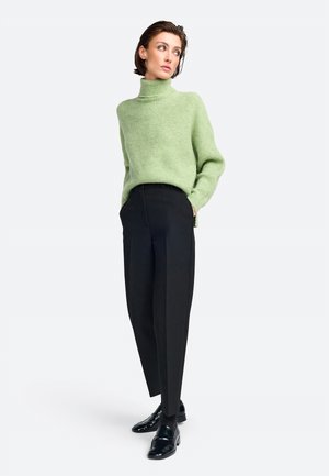 Grüner, strukturierter Rollkragenpullover kombiniert mit schwarzen maßgeschneiderten Hosen und glänzenden schwarzen Slippern. Schlichte, klare Linien dominieren das Outfit.