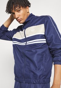 Navy track jacket made of smooth fabric, featuring a beige panel with two white stripes. Includes zip closure and elastic cuffs.