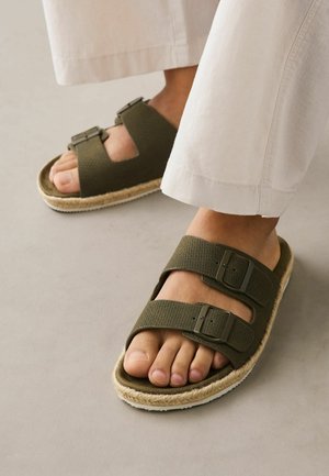 Füße, die olivgrüne Sandalen mit Doppel-Schnalle, strukturierten Riemen und geflochtenem Sohlenbesatz tragen, kombiniert mit cremefarbenen, weiten Hosen.