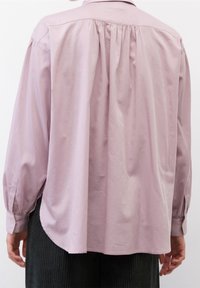 Camicia a maniche lunghe di un morbido rosa, con yoke posteriore con pieghe, polsini con bottone e un orlo curvo. Indossata con pantaloni neri a trama.