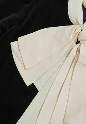 Tkanina v krémové barvě s texturovaným povrchem, zdobená velkou mašlí. Umístěná na jednobarevném černém pozadí.