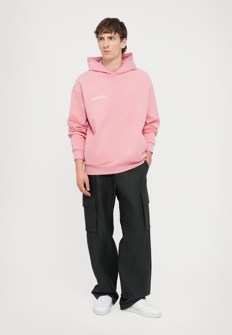 Sweatshirt à capuche rose avec un motif à l'avant et un pantalon cargo large noir. Des baskets blanches complètent l'ensemble. Fond blanc uni.