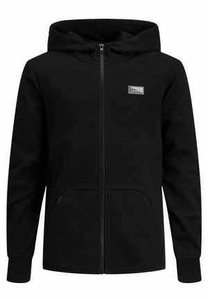 Persona vistiendo unos jeans azul claro, zapatillas blancas, camiseta gráfica blanca y sudadera negra con cremallera, con las manos en los bolsillos sobre un fondo gris.