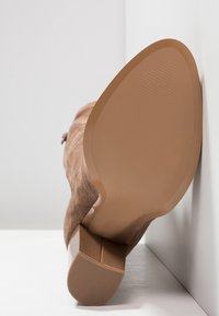 Botte en daim marron reposant semelle vers le haut contre un mur blanc, montrant une semelle lisse et un talon carré avec une surface texturée antidérapante.