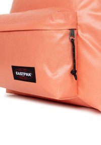 Persikofärgad Eastpak-ryggsäck med en slät, strukturerad ytfinish, som har en svart logopatch och en dragkedja på framfickan.