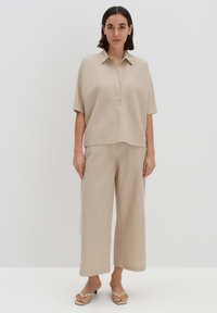 Polo shirt beige a maniche corte con placket a bottoni, abbinato a pantaloni beige a gamba larga. Tessuto morbido con una vestibilità rilassata, indossato con sandali.