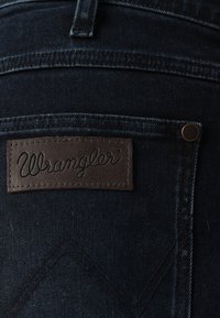 Tamno plavi traperice s smešnim kožnim detaljem s utisnutim natpisom "Wrangler." Sadrži bakrene zakovice i suptilne šavovske uzorke.