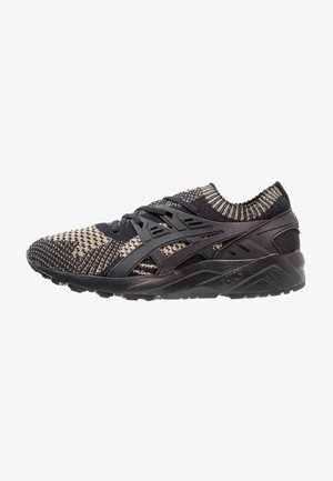 Zwart-beige gebreide hardloopschoen met zwarte vetersteuncage en dikke zwarte zool, zijaanzicht van links.