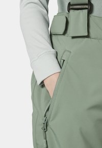 Los pantalones verde claro cuentan con un bolsillo con cremallera y una correa vertical con hebilla. La tela es suave y de textura agradable, llevándose encima de una blusa gris.