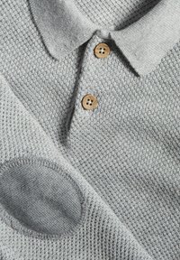 Gråstickad polo tröja med en strukturerad väv, tre träknappar och en cirkulär patchdetalj på ärmen.