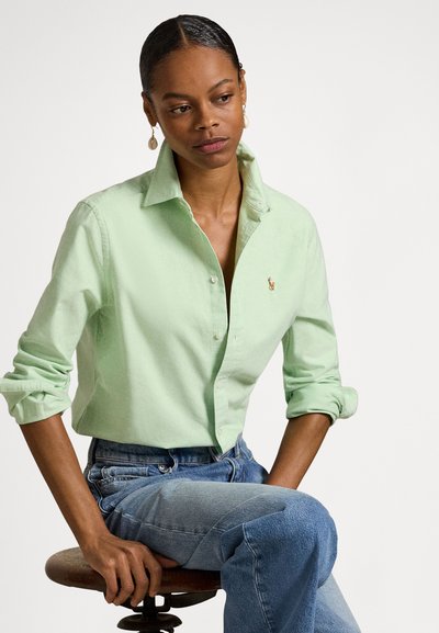 Gaismas zaļa kokvilnas krekls ar pogu aizdares apkakli, uzrullētām piedurknēm un nelielu izšūtu logo. Kopā ar augsta vidukļa džinsa biksēm.