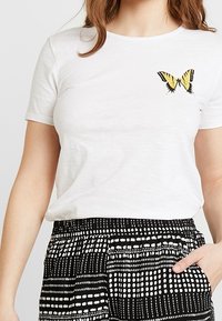 Camiseta blanca de manga corta con una mariposa bordada en amarillo y negro en el pecho izquierdo. Combinada con pantalones negros con estampado.