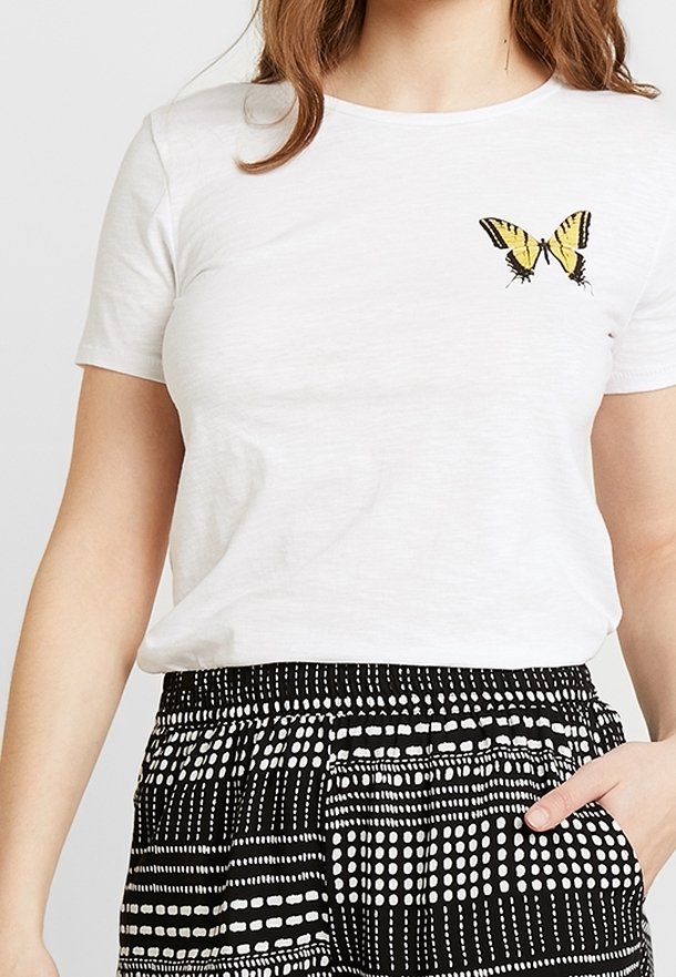 Camiseta blanca de manga corta con una mariposa bordada en amarillo y negro en el pecho izquierdo. Combinada con pantalones negros con estampado.