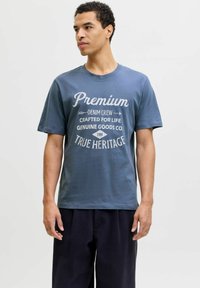 Man som bär en blå t-shirt med vit text "Premium Denim Crew Crafted for Life Genuine Goods Co. True Heritage" som står mot en enfärgad bakgrund.