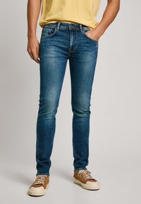 Jeans in denim blu con vestibilità slim, tonalità sbiadite, cinque tasche e cuciture a contrasto, abbinati a sneakers marroni.