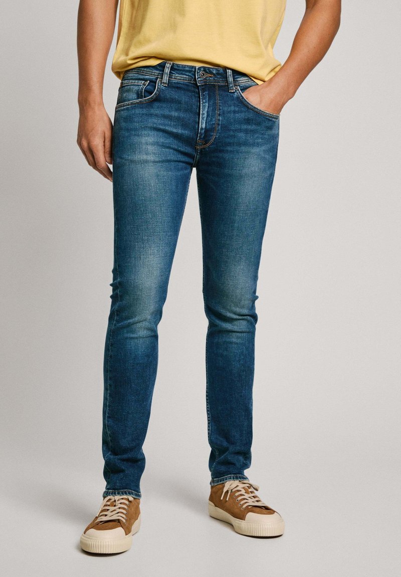Jeans in denim blu con vestibilità slim, tonalità sbiadite, cinque tasche e cuciture a contrasto, abbinati a sneakers marroni.