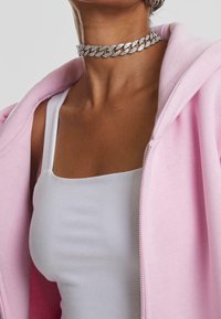 Sudadera rosa con cremallera y textura suave, usada sobre una camiseta blanca sin mangas. Lleva un collar gargantilla de cadena plateada gruesa con detalles brillantes.