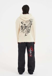 Rippender cremefarbener Hoodie mit einem großen schwarzen Grafikdesign, das einen Tiger und einen Schädel auf dem Rücken zeigt; kombiniert mit dunklen, locker sitzenden Jeans mit roten Motiven.