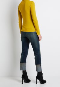 Gelbes Langarmoberteil kombiniert mit eng anliegenden dunkelblauen Jeans, die an den Knöcheln aufgerollt sind, und schwarzen, hochhackigen Ankle-Boots mit elegantem Design.