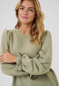 Roheline tekstuuriga pluus puffesleeved ja kogutud mansettidega. Ümmargune kaelus, kerge kangas ja lõdvestatud lõige.