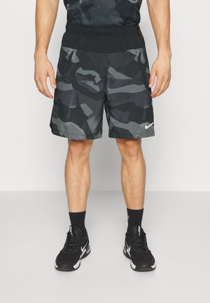 Mężczyzna noszący czarne i szare sportowe szorty w kamuflażowy wzór, czarne skarpety i czarne buty sportowe Nike stoi na jasnej podłodze.