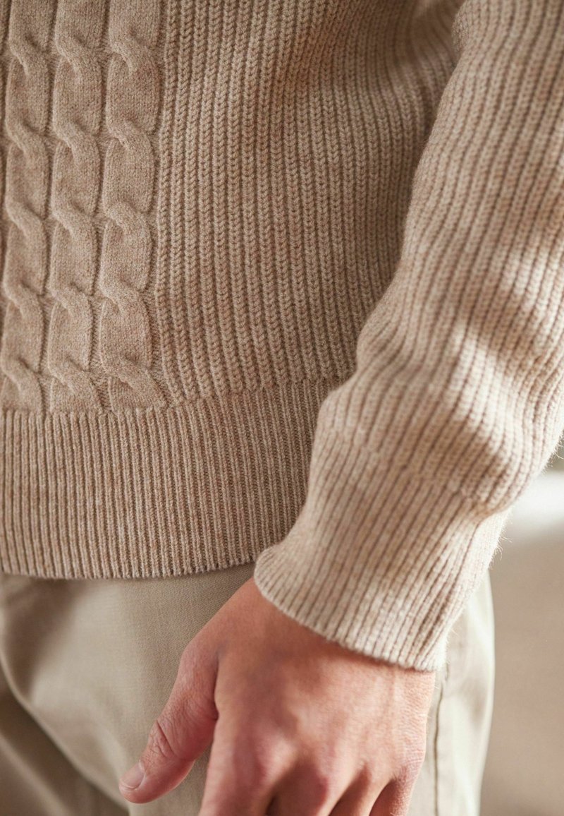 Suéter de punto beige con un patrón texturizado de cable en el lado izquierdo y puños acanalados, mostrado junto a una mano sobre una tela beige.