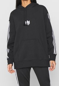 Black hoodie with a front pocket, white floral logo, and white vertical stripes on sleeves. Made from soft fabric, relaxed fit.