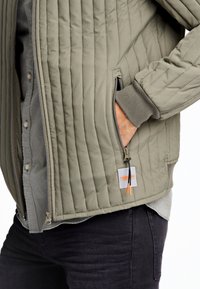 Chaqueta acolchada verde con un bolsillo con cremallera, puños grises y un acento naranja en la cremallera. Presenta un cierre frontal con botones y un forro interior.