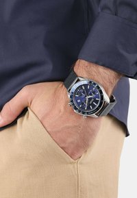 Silberfarbene Armbanduhr mit blauem Zifferblatt, Mesh-Armband, Chronographenfunktion und Datumsanzeige. Die Hand liegt in einer Tasche von beigen Hosen.