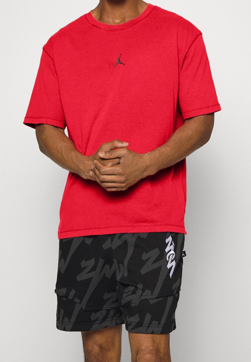 Camiseta de algodón roja con un logo negro de Jumpman en el pecho, combinada con pantalones cortos negros que presentan un patrón abstracto gris y acentos blancos.