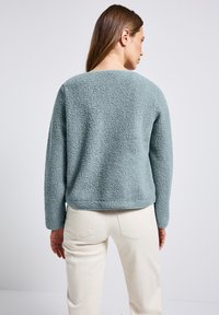 Maglione fuzzy color teal con scollatura a girocollo e maniche lunghe, caratterizzato da una superficie testurizzata e una vestibilità comoda, abbinato a pantaloni chiari.