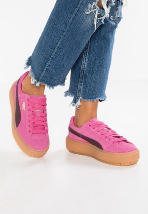 Person iført lyserøde ruskindssneakers med gummisåler og sorte striber, kombineret med blå denimjeans med frynsede kanter på en hvid gulv.