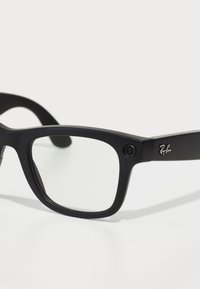 Occhiali da sole Ray-Ban con montatura in plastica nera opaca, forma rettangolare, lenti trasparenti e una piccola fotocamera sul lato. Logo sulla tempia.