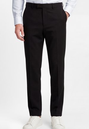 Mann trägt schlanke schwarze Anzughose, weißes Hemd und weiße Sneakers, steht mit einer Hand in der Tasche.