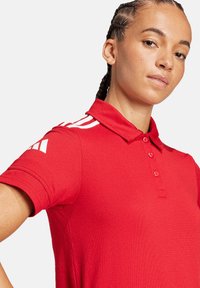 Polo shirt rossa realizzata in tessuto testurizzato, con colletto, patta con tre bottoni e dettagli a strisce bianche sulle spalle.