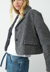 Skórzana szara kurtka z wyciętym kołnierzem, dwoma czarnymi guzikami i kieszeniami z przodu, w zestawie z białą koszulą i niebieskimi dżinsami.