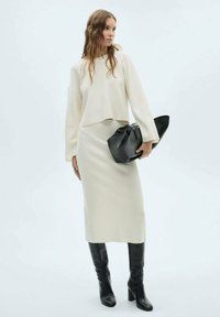Ensemble en tricot crème, comprenant un pull et une jupe, complété par des bottes noires hautes et un sac à main noir structuré. Texture lisse, coupe moderne.