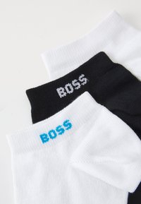 Trois paires de chaussettes de cheville : noires avec logo blanc, blanches avec logo bleu, toutes fabriquées en tissu lisse et respirant, avec des bords côtelés.