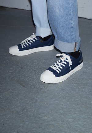 Person iført marineblå og hvide sneakers med hvide snørebånd, sorte sokker og lyseblå opsmøgede jeans, der står på en grå gulv.