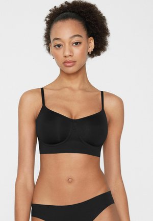 Jeune femme aux cheveux bouclés portant un soutien-gorge noir et une culotte assortie, debout devant un fond blanc uni.