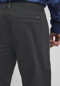 Pantaloni negri dintr-un material cu textură netedă, având un buzunar posterior cu închidere prin buton și o etichetă de marcă.