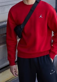 Sudadera roja con cuello redondo y logo negro de Jordan en el pecho. Combinada con pantalones jogger negros que tienen un zipper y detalles de logo en blanco.