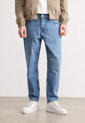 Homme portant un jean bleu clair, des baskets blanches, une veste beige, debout sur un sol gris devant un fond blanc uni.