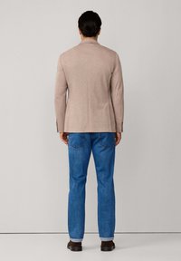 Hellbeige Blazer mit strukturierten Schultern, kombiniert mit blauen Jeans, die zwei Gesäßtaschen und hochgekrempelte Bündchen haben, getragen mit braunen Schuhen.
