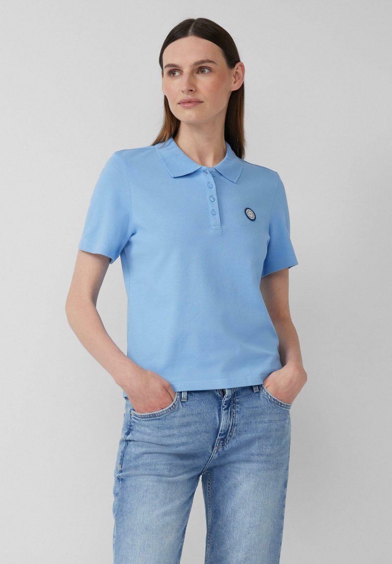 Hellblaues Polohemd aus Baumwolle, mit Kragen, kurzen Ärmeln und einer Knopfleiste sowie einem runden Logopatch auf der linken Brust.