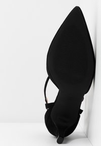 Chaussure à talons pour femmes à bout pointu noir, montrant une semelle texturée et un talon aigu étroit sur fond blanc.