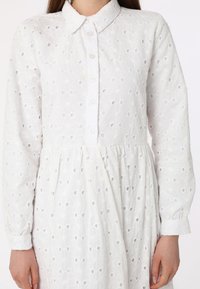 White embroidered dress featuring floral patterns, long sleeves with buttoned cuffs, a collar, and a gathered waistline for a relaxed fit.
