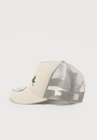 Μπεζ καπέλο με κυρτή γείσο, πλέγμα στο πίσω μέρος και κεντημένο πράσινο φύλλο στο μπροστινό μέρος. Διαθέτει κούμπωμα τύπου snapback για ρυθμιζόμενο μέγεθος.