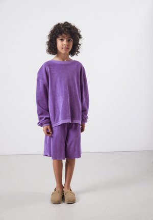 Dieťa s kučeravými vlasmi, oblečené v voľnej fialovej mikine a zodpovedajúcich šortkách, stojace na svetlej podlahe pred jednoduchou bielou stenou.